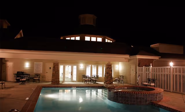 Night view of an outdoor swimming pool and hot tub area at a senior living facility, with patio chairs and tables arranged around the pool. The building behind has illuminated windows and stone pillars, and the area is enclosed by a white fence.