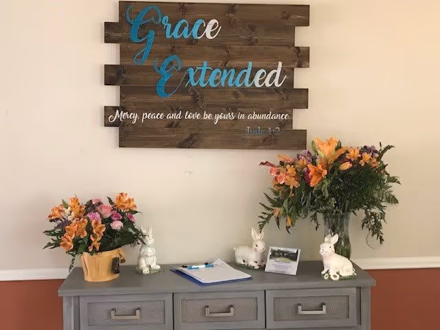 A decorative interior wall display featuring a wooden sign with the words 'Grace Extended' and a Bible verse from Jude 1:2. Below the sign is a gray table with three drawers, adorned with two flower arrangements in vases, several white ceramic bunny figurines, a clipboard with paper and pen, and a small framed photo.