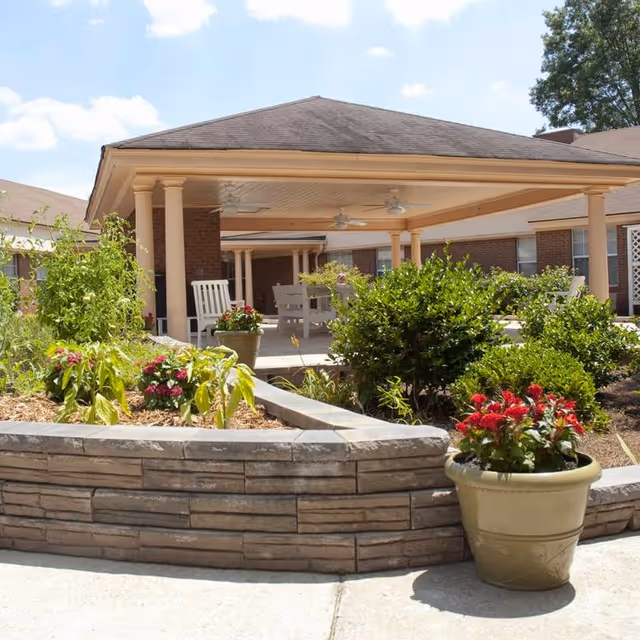 Outdoor patio area at Southland Health & Rehabilitation featuring a covered seating area with white chairs and ceiling fans, surrounded by landscaped garden beds with green shrubs and colorful flowers in pots and soil beds.