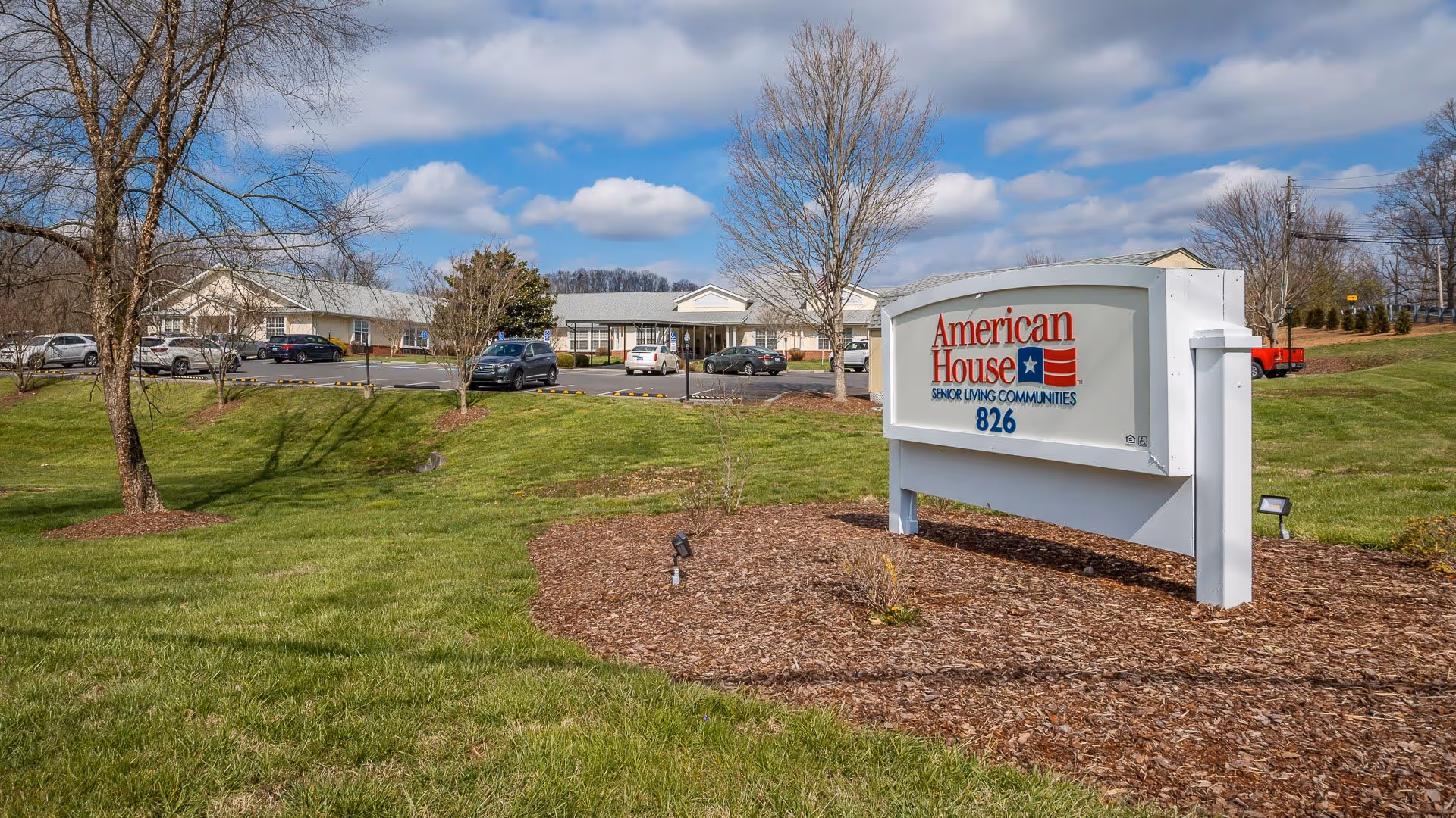 Outdoor view of American House Bristol senior living community with a large white sign displaying the facility name and address number 826, surrounded by green grass, trees, and a parking lot with several cars under a partly cloudy sky.