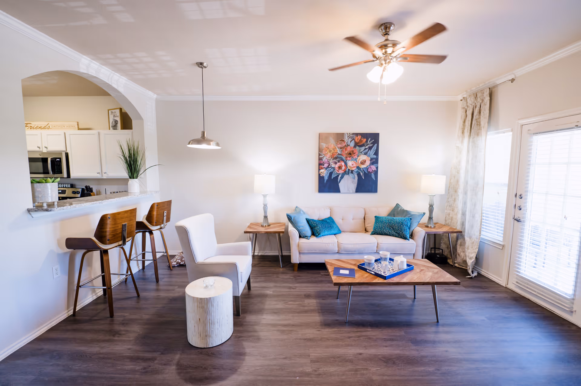 A bright and modern living room with a beige sofa adorned with teal pillows, two wooden side tables with lamps, a wooden coffee table with a blue tray and cups, a white armchair, and two wooden bar stools at a kitchen counter. The room features a ceiling fan with lights, a large window with curtains, and a colorful floral painting on the wall.