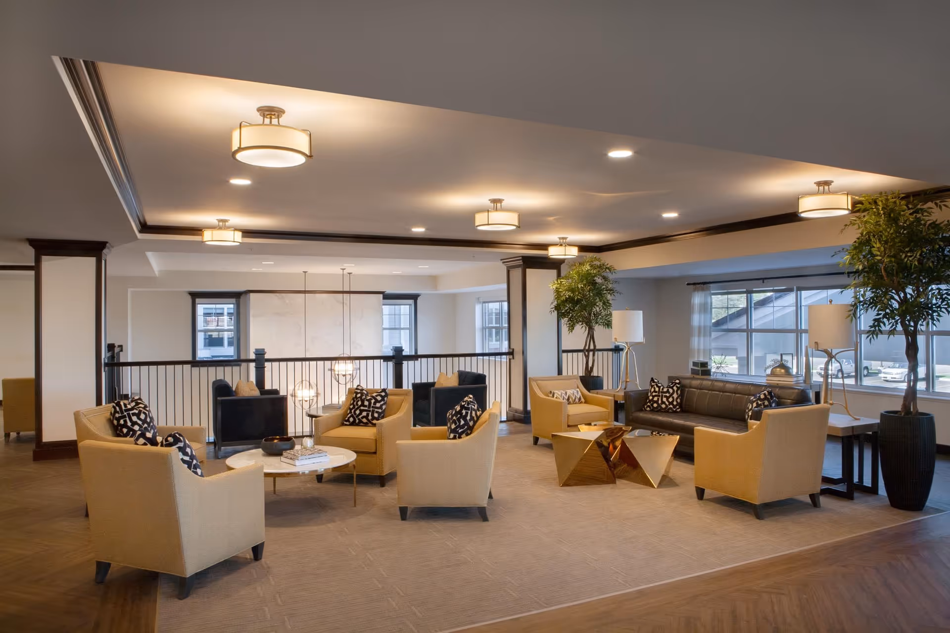 A spacious and well-lit living room area in a senior living facility with multiple beige armchairs and a black leather sofa arranged around two modern coffee tables. The room features large windows, potted plants, floor lamps, and ceiling lights, creating a warm and inviting atmosphere.