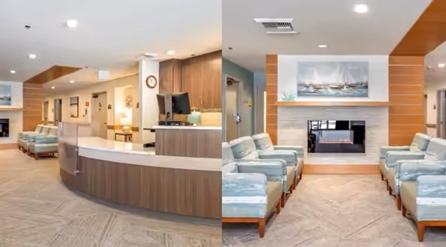 Interior view of a senior living facility reception area and lounge. The reception desk is curved with a computer and decorative items on it. The lounge area features multiple cushioned chairs arranged facing a modern fireplace with a painting of sailboats above it. The space is well-lit with recessed ceiling lights and has a neutral color palette with wood accents.