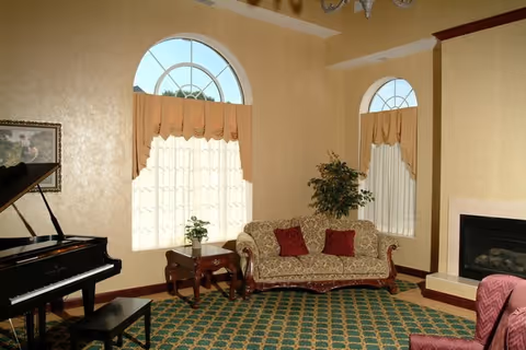 A cozy living room with a patterned green carpet, a grand piano with a bench, a floral upholstered sofa with red cushions, a wooden side table with a small plant, a fireplace, and two large arched windows with beige curtains.