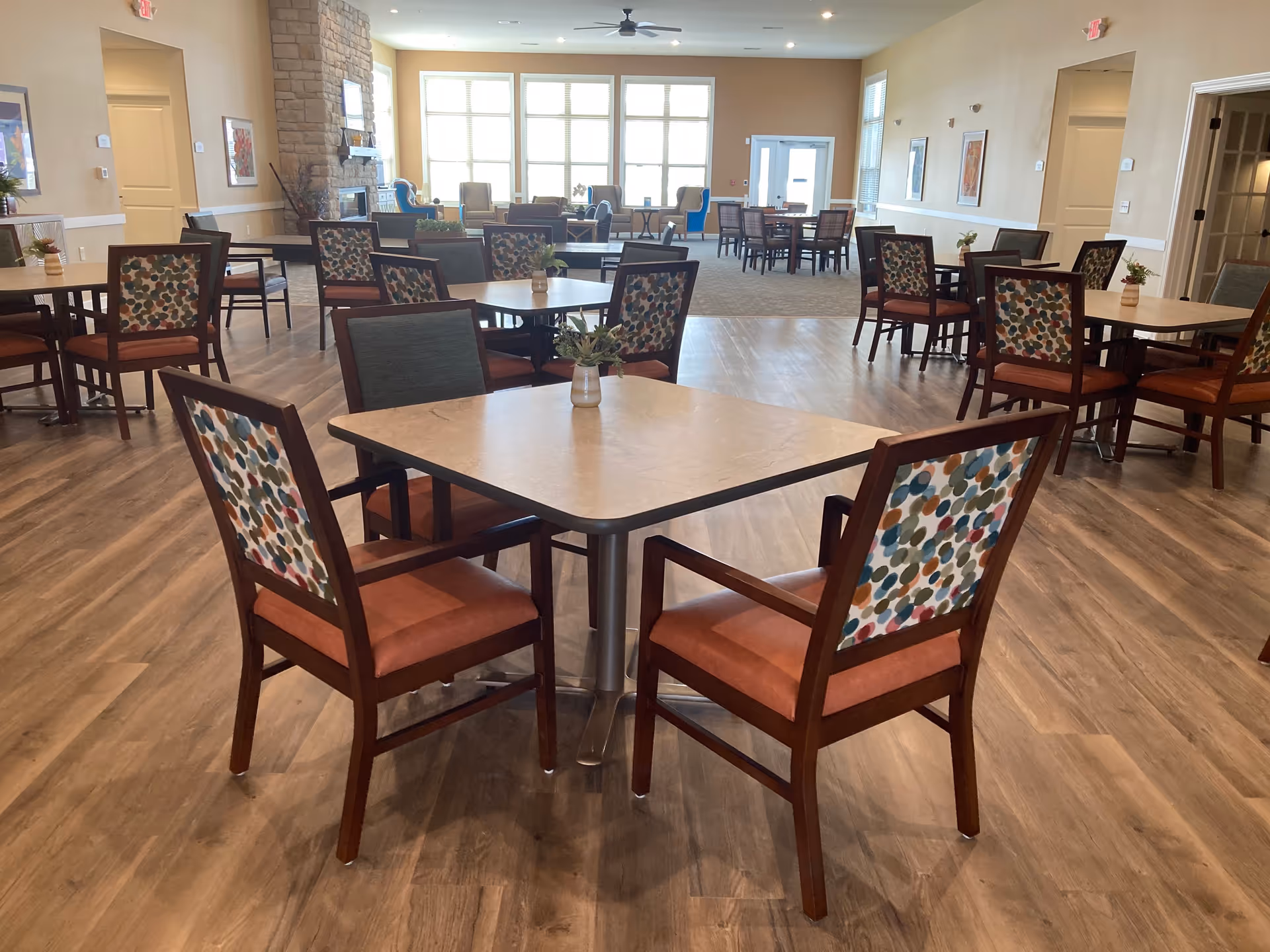 A spacious dining room with multiple tables and chairs arranged neatly. The chairs have wooden frames with patterned backs and orange cushions. The room has large windows letting in natural light, a stone fireplace on the left, and a ceiling fan. The floor is a combination of wood and carpeted areas, and there are framed pictures on the walls.