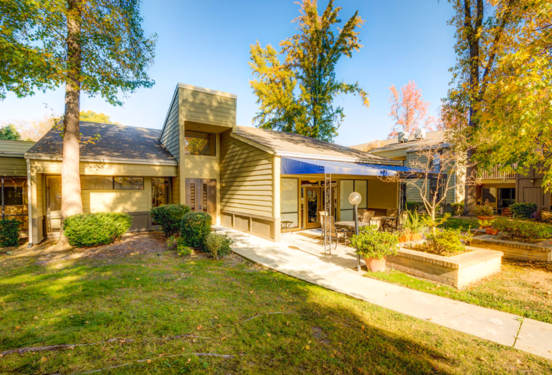 Sunlit courtyard and main entrance of a senior living building with outdoor seating, a walkway, and mature trees.