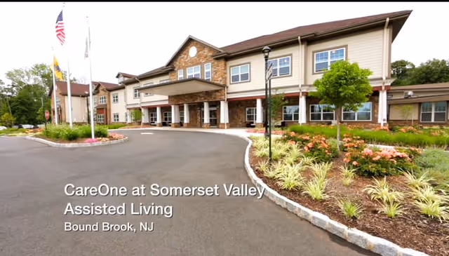 Exterior view of CareOne at Somerset Valley Assisted Living facility in Bound Brook, NJ, showing a two-story building with a covered entrance, landscaped garden with flowers and shrubs, and three flagpoles with flags.