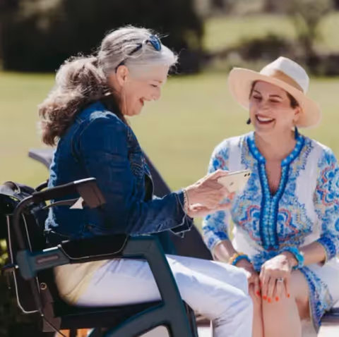 Two women outdoors enjoying a sunny day, one sitting in a wheelchair and showing something on her phone to the other woman who is wearing a hat and a patterned dress. Both are smiling and appear to be engaged in a pleasant conversation.
