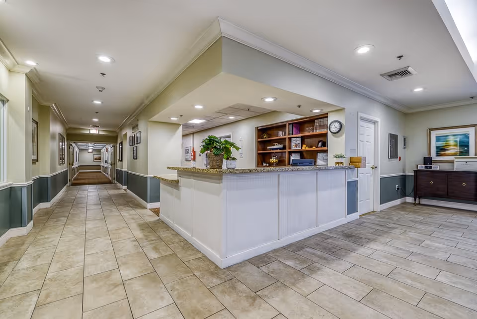 Interior view of a senior living facility hallway with a reception desk on the right side. The reception desk has a granite countertop with a potted plant and a clock on the wall behind it. The hallway is well-lit with ceiling lights and has framed pictures on the walls. There is a wooden cabinet with a printer and framed artwork on the far right side.