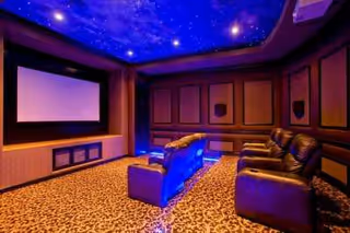 A luxurious home theater room with a large projection screen, leather recliner chairs arranged in two rows, dark paneled walls, and a ceiling designed to look like a starry night sky with blue lighting.