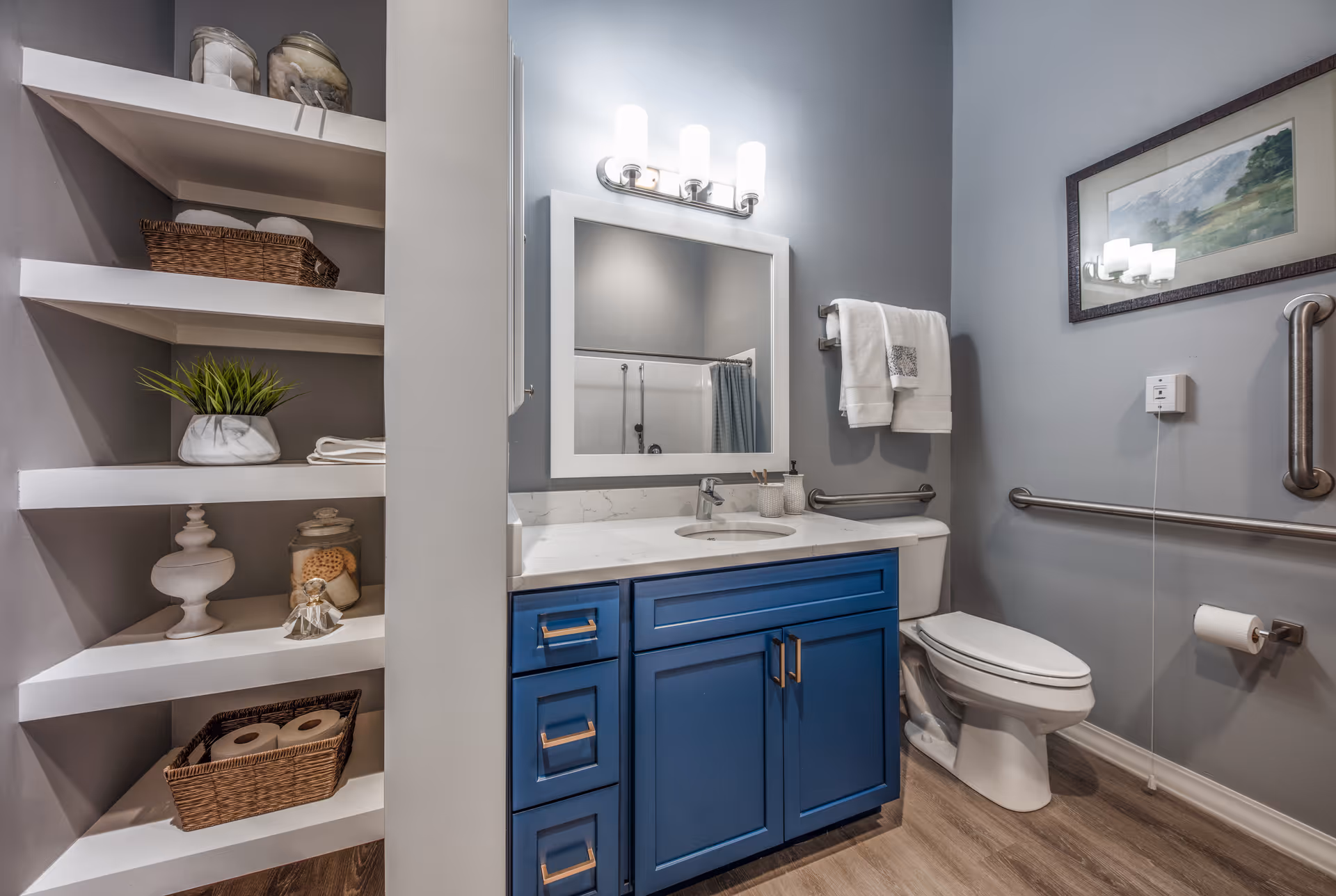 A modern bathroom with a blue vanity cabinet, white countertop, and an oval sink. Above the sink is a rectangular mirror with three light fixtures. To the right is a white toilet with grab bars on the wall and a towel rack holding two white towels. On the left side, there are built-in white shelves holding decorative items, towels, and toilet paper. The walls are painted light gray, and there is a framed landscape painting above the toilet.