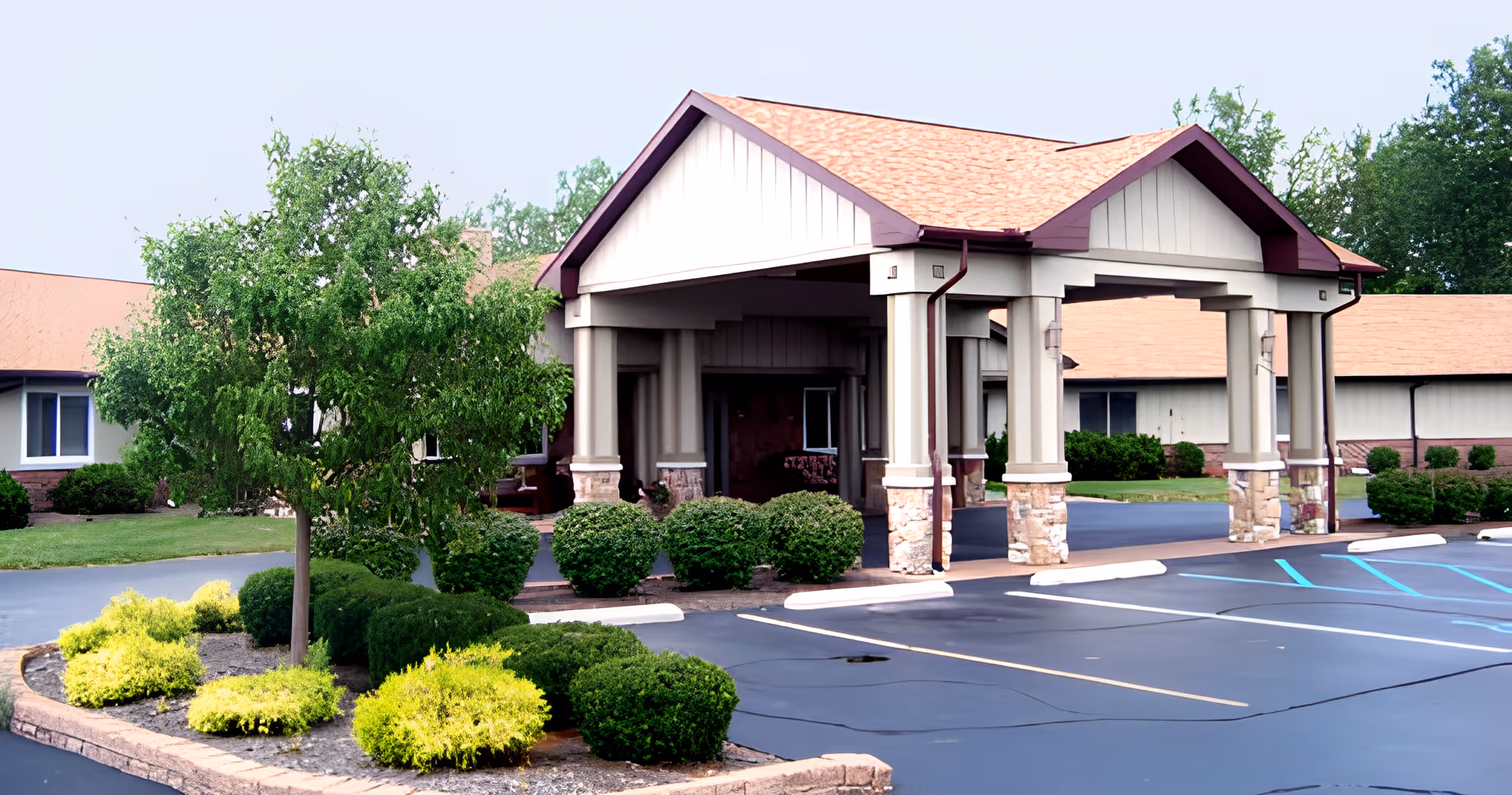 Exterior view of Briar Hill Health Campus - Memory Care showing the entrance with a covered drop-off area supported by columns. The building has a beige facade with a reddish-brown roof. There are neatly trimmed bushes and a small tree in landscaped areas near the entrance, and an empty parking lot with marked spaces in front.