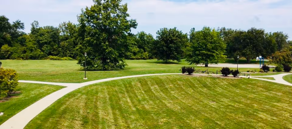 A well-maintained outdoor grassy area with curved concrete walking paths, several trees, bushes, and a basketball hoop in the distance under a partly cloudy sky.