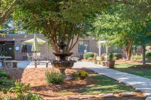 Brick senior living facility courtyard with fountain, benches, trees, and shaded seating.