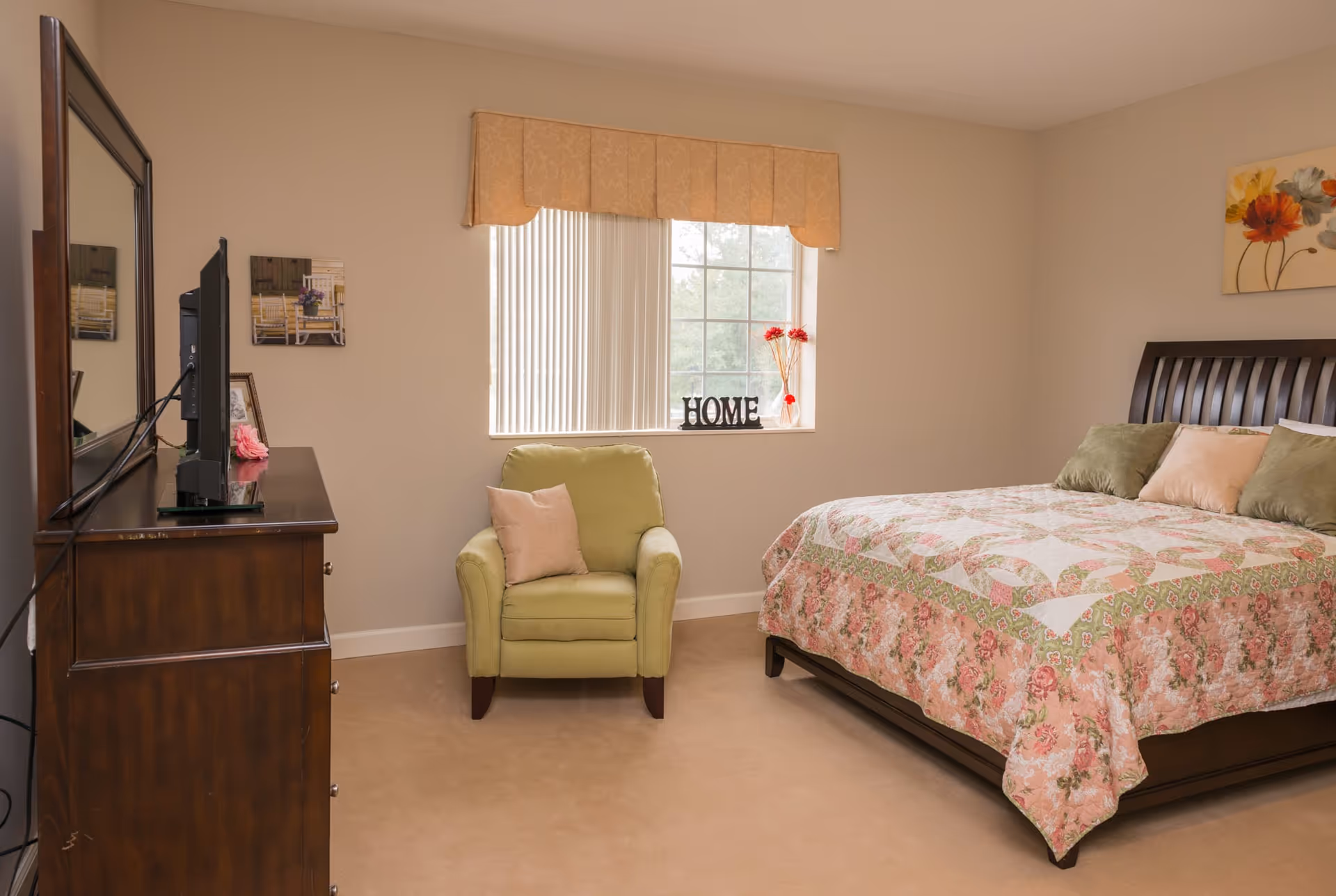 A cozy bedroom with a wooden bed frame and a floral quilted bedspread, decorated with green and beige pillows. Next to the bed is a green armchair with a beige pillow. A window with vertical blinds and a beige valance lets in natural light. On the windowsill, there is a decorative 'HOME' sign and a vase with red flowers. A wooden dresser with a TV on top is positioned opposite the bed. The walls are painted beige and decorated with framed pictures.
