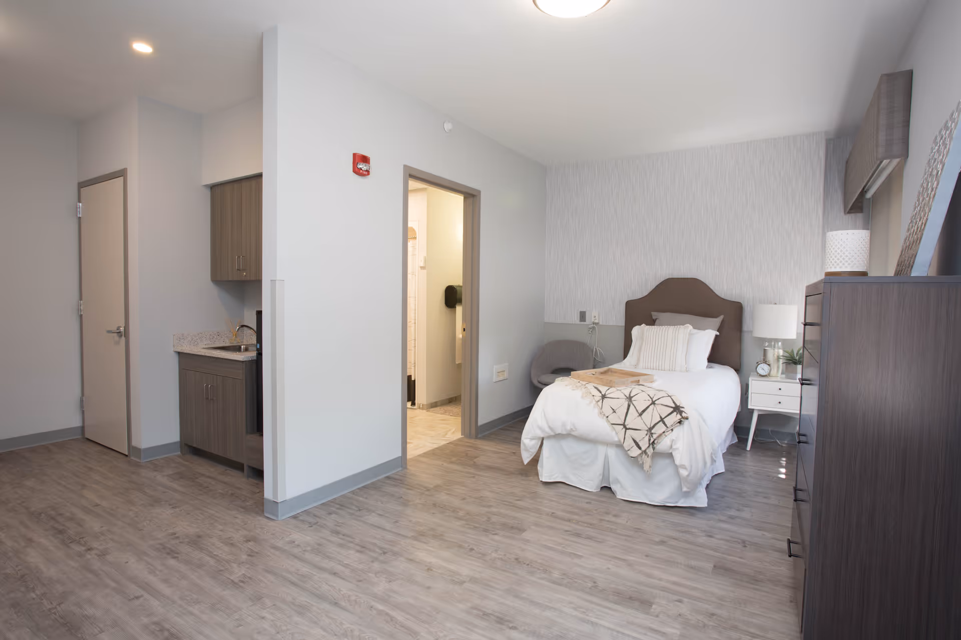 A clean and modern bedroom in a senior living facility featuring a single bed with white bedding and a decorative throw, a nightstand with a lamp and clock, a gray armchair, a tall dresser, and a small kitchenette area with cabinets and a sink. The room has light gray walls and wood-look flooring, with an open doorway leading to a bathroom.