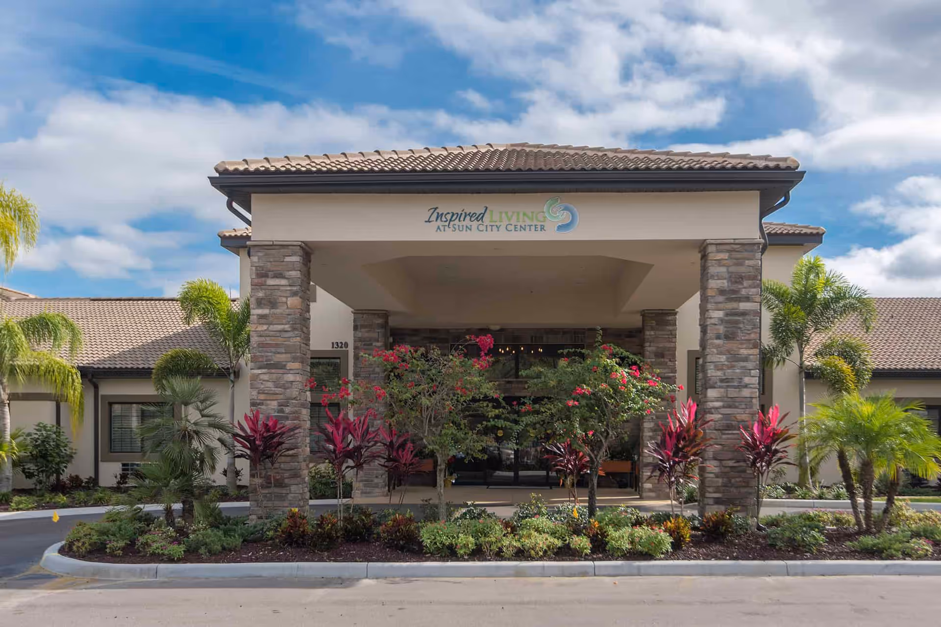 Front entrance of a single-story senior living facility with stone pillars, landscaped plants, and a sign reading "Inspired Living at Sun City Center".