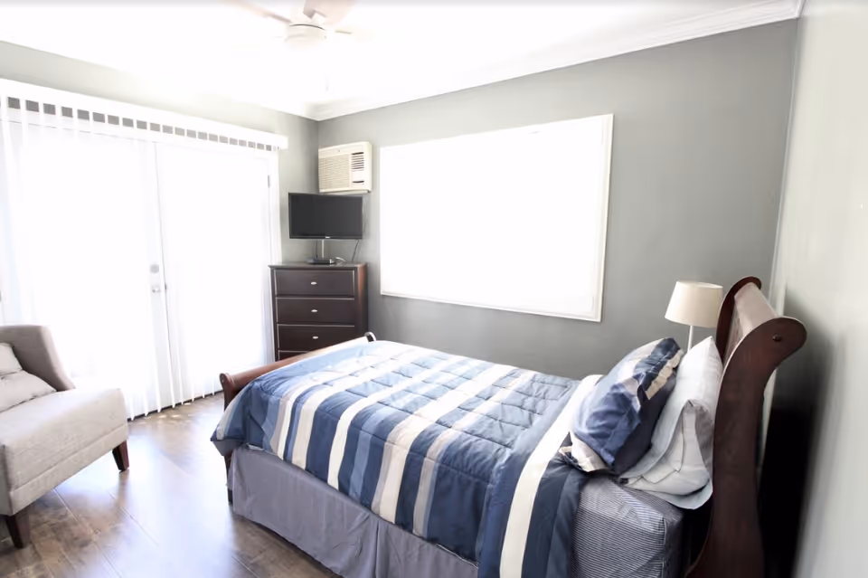 Bright bedroom with a wooden bed dressed in blue-striped bedding, a dresser with a small TV, an armchair, and sliding glass doors.