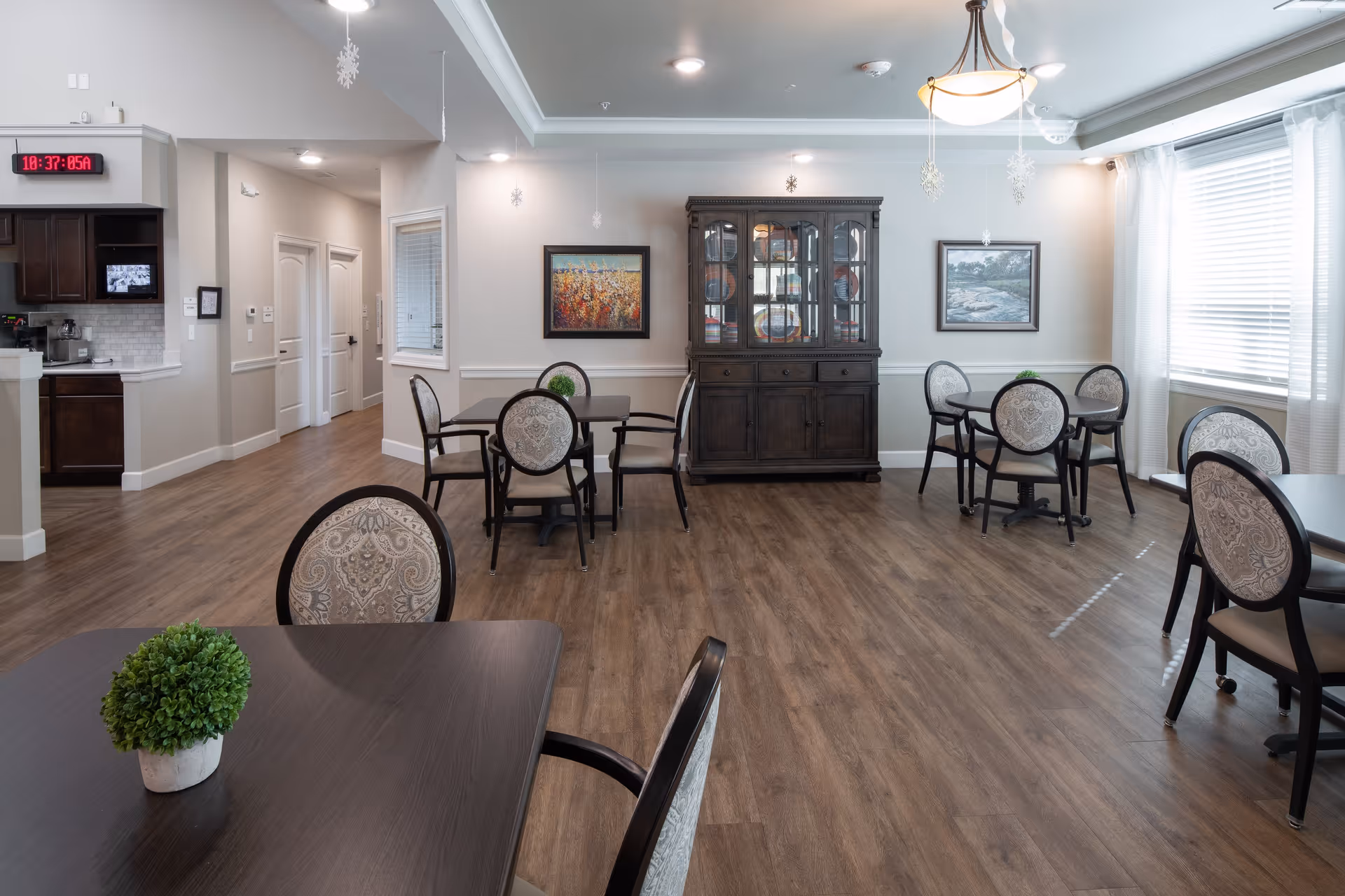 A bright and spacious dining room in an assisted living facility with several dark wood tables and upholstered chairs arranged neatly. The room features wood flooring, light-colored walls with two framed paintings, a dark wood china cabinet, and large windows with white curtains allowing natural light to fill the space. A small green plant is placed on the table in the foreground. The kitchen area is partially visible to the left, and ceiling lights and hanging snowflake decorations are present.