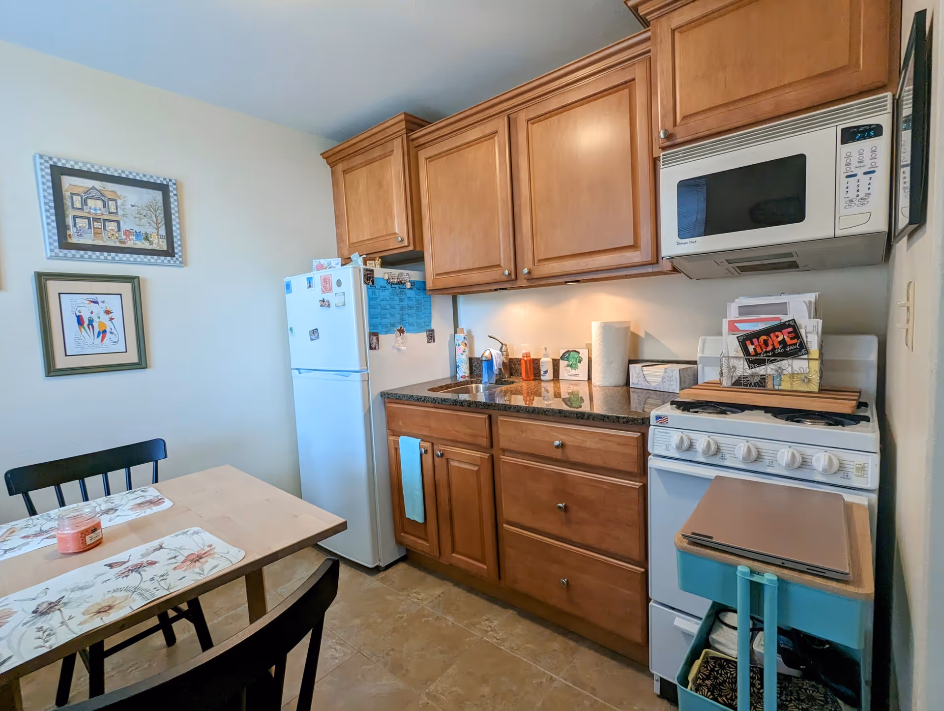 A small kitchen area with wooden cabinets, a white refrigerator, a white stove with a microwave above it, and a granite countertop with a sink. There is a small dining table with two black chairs and floral placemats. Two framed pictures hang on the wall above the refrigerator.