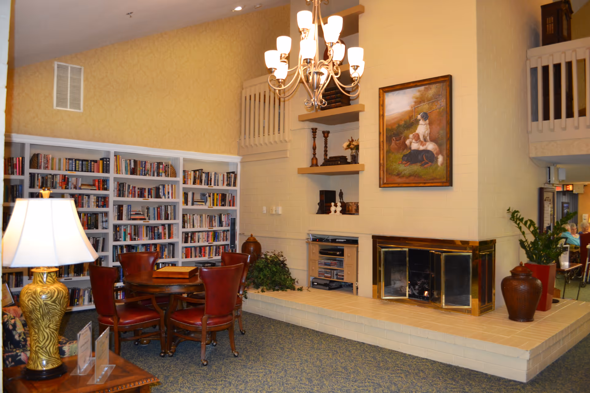 A cozy communal lounge with bookshelves, a round table and red chairs, a fireplace with a painting above, and a chandelier.