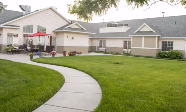 Curved concrete path through a green lawn leading to a single-story assisted living building with outdoor seating and a red patio umbrella.