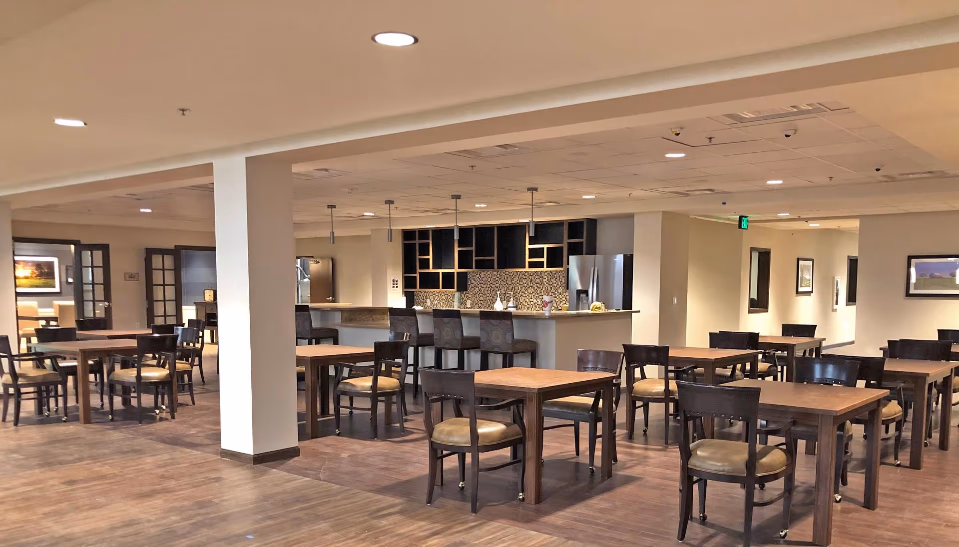 A spacious dining area with multiple wooden tables and chairs arranged neatly. In the background, there is a kitchen area with a counter, bar stools, and modern appliances. The room has a warm, inviting atmosphere with soft lighting and framed pictures on the walls.