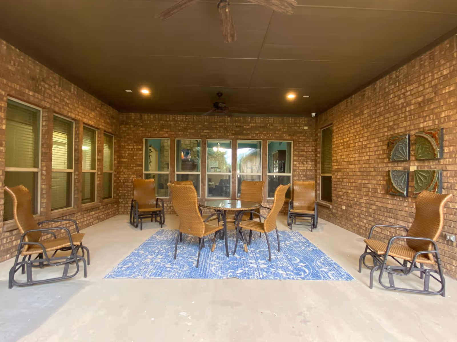 Covered outdoor patio area with six wicker rocking chairs arranged around a round glass table on a blue patterned rug. The walls are made of brown brick with multiple windows and decorative wall art. The ceiling is dark with recessed lighting and a ceiling fan.