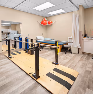 Physical therapy room with a wooden ramp and black handrails for walking exercises, an examination table with a pillow, red exercise balls stored on a wall-mounted rack, a stool, and medical equipment on a counter with a curtain divider.