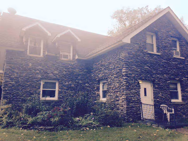 Two-story stone-clad house with dormer windows, a white front door, and a small garden and lawn.