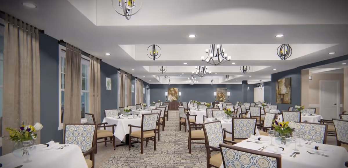 A spacious dining room with multiple tables covered in white tablecloths, each set with silverware, napkins, and floral centerpieces. The room features patterned carpet, blue walls, large windows with beige curtains, and modern chandeliers hanging from the ceiling.