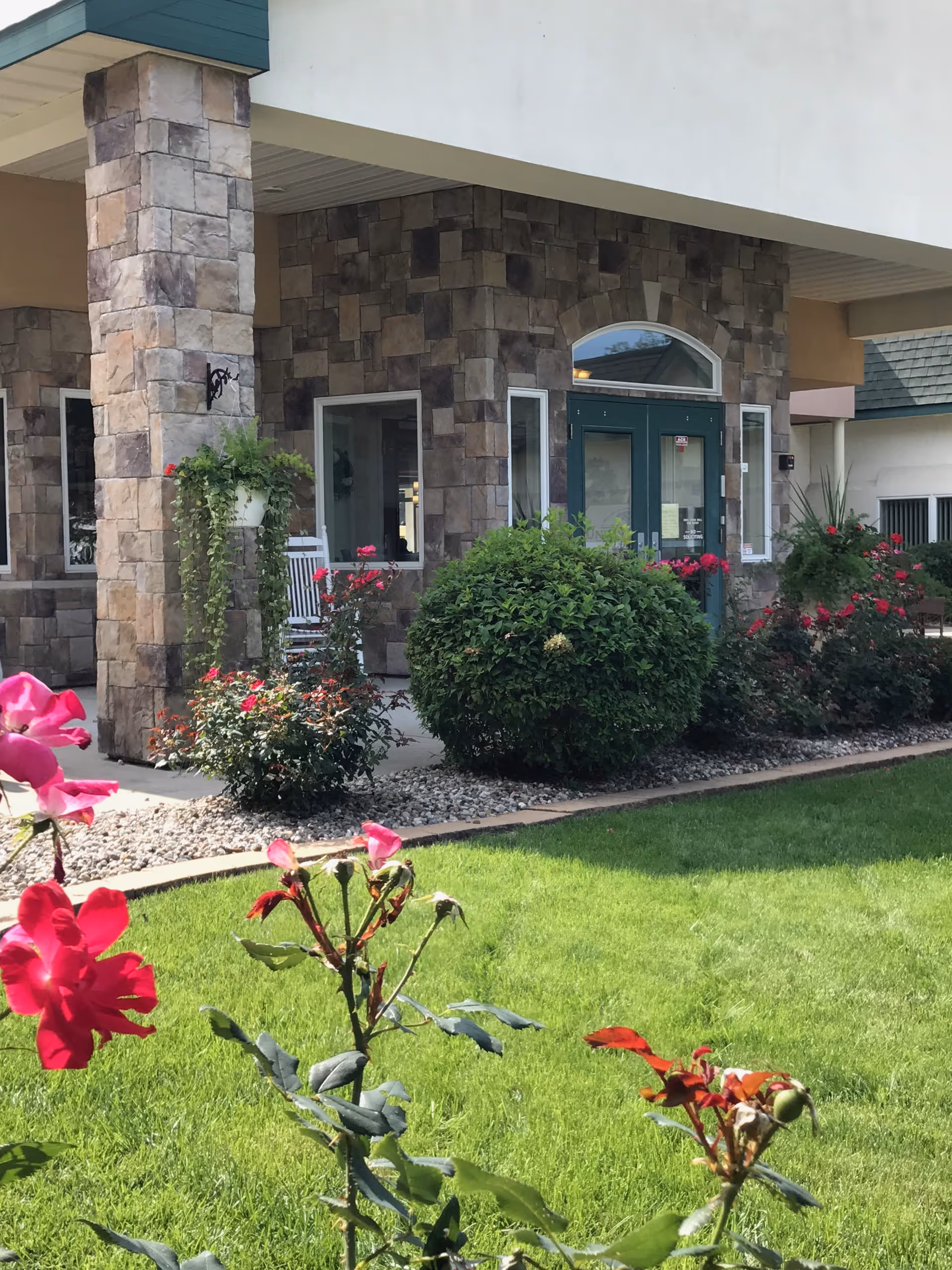 Front entrance of a stone-clad assisted living building with green double doors, columns, a porch and flowering shrubs on a manicured lawn.