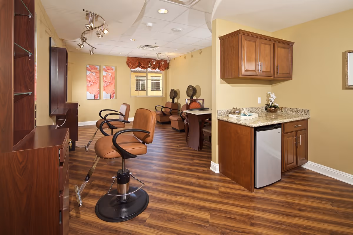 Interior view of a salon area in a senior living facility with wooden flooring, brown salon chairs, hair drying stations, a small kitchenette with granite countertop and wooden cabinets, and wall art with floral designs.