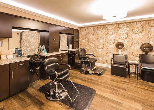 Interior of a salon room with two black salon chairs in front of mirrors and sinks, a patterned wallpaper on one wall, wooden flooring, and two hair dryer chairs with hooded dryers.