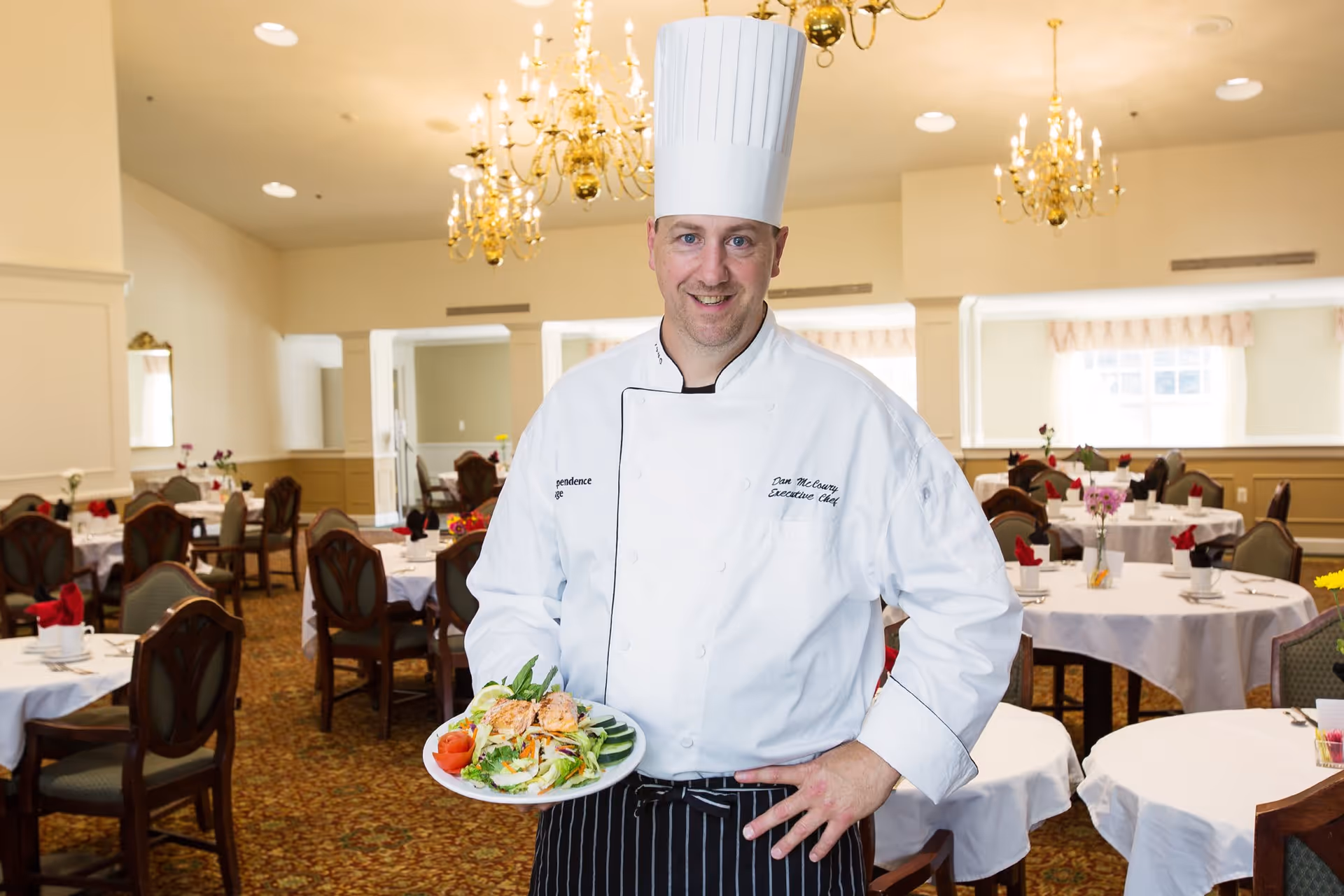 A chef wearing a white chef's coat and tall white hat stands in a dining room holding a plate of salad with grilled salmon. The dining room has round tables covered with white tablecloths, set with plates, glasses, and red napkins. The room is decorated with chandeliers and has a warm, inviting atmosphere.