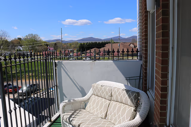 A small balcony with a cushioned wicker loveseat, black metal railing, and a view of trees, rooftops, and distant mountains under a clear blue sky.