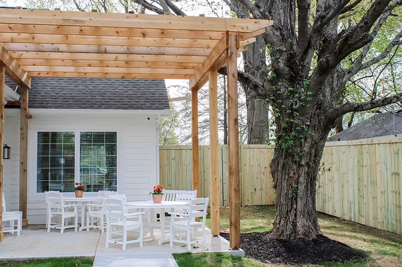 Outdoor patio area with white tables and chairs under a wooden pergola next to a large tree and a wooden fence surrounding the yard.