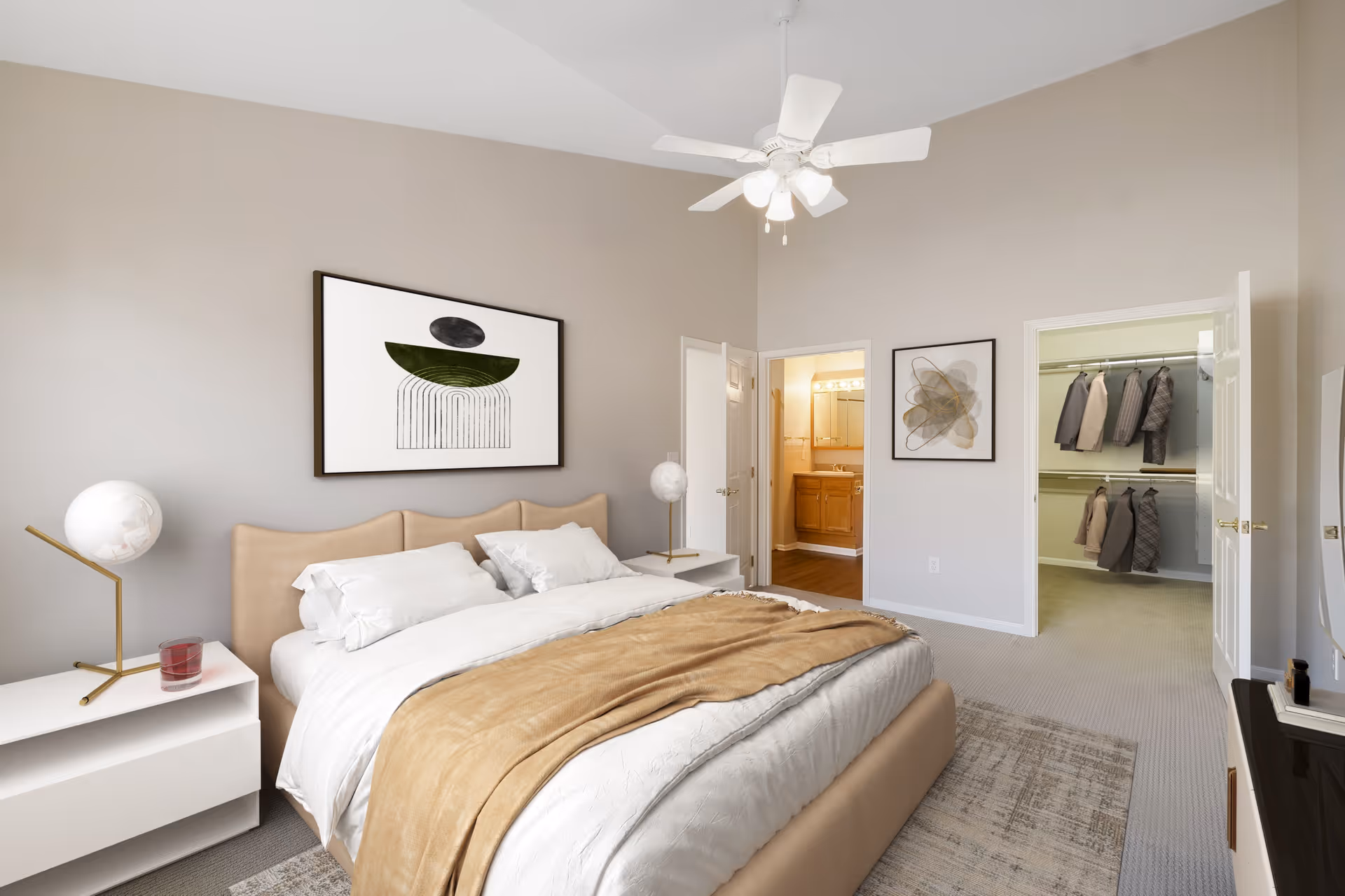 A modern bedroom with a large bed featuring white bedding and a beige throw blanket. There are two white nightstands with spherical lamps on each side of the bed. The walls are painted light beige and decorated with abstract artwork. The room has a ceiling fan with lights. There are two open doors, one leading to a bathroom with a wooden vanity and the other to a walk-in closet with hanging clothes.