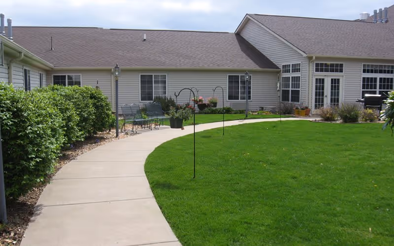 A curved concrete walkway runs through a well-maintained green lawn with garden hooks and a few benches along the path. The walkway leads to a single-story building with beige siding, multiple windows, and a glass door entrance. There are bushes and plants along the left side of the walkway and near the building.