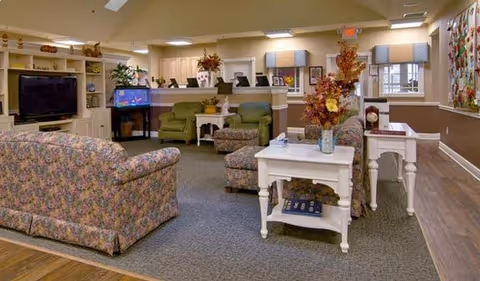 Cozy senior living lounge with floral upholstered sofas, white side tables, a television and fish tank, and a reception desk in the background.