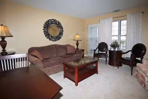 A cozy living room with a brown sofa, two dark wooden chairs with round backs, a glass-top coffee table, a side table with a lamp, and a large decorative round mirror on the wall. There is a window with white curtains letting in natural light.