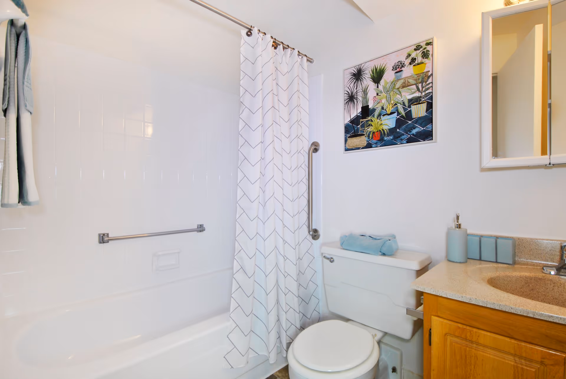 A clean bathroom featuring a white bathtub with a white shower curtain that has a subtle chevron pattern. There is a silver grab bar on the bathtub wall and a towel hanging on the left side. Next to the bathtub is a white toilet with a folded blue towel on top. Above the toilet is a colorful framed picture of various potted plants. To the right is a wooden vanity with a beige countertop, a soap dispenser, and a mirrored medicine cabinet on the wall.