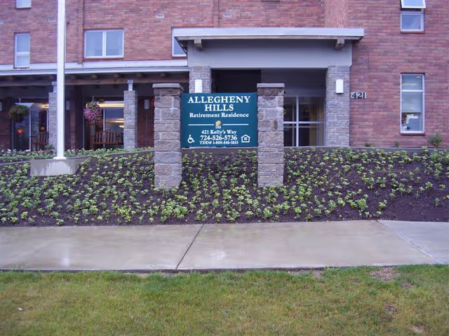 Front exterior view of Allegheny Hills Retirement Residence showing a brick building with a covered entrance, a green sign with white text displaying the facility name, address, and contact information, surrounded by a landscaped area with small plants and a concrete sidewalk in front.