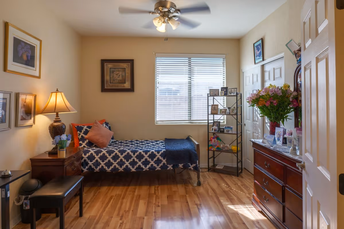 A cozy bedroom with a single bed covered in a blue and white patterned bedspread and orange and pink pillows. There is a wooden nightstand with a lamp and a small potted plant next to the bed. The room has wooden flooring, a ceiling fan with lights, and a window with blinds letting in natural light. On the right side, there is a wooden dresser with a large bouquet of flowers and various framed photos and cards. A black metal shelving unit with more framed photos and small items stands near the window. The walls are decorated with framed artwork.