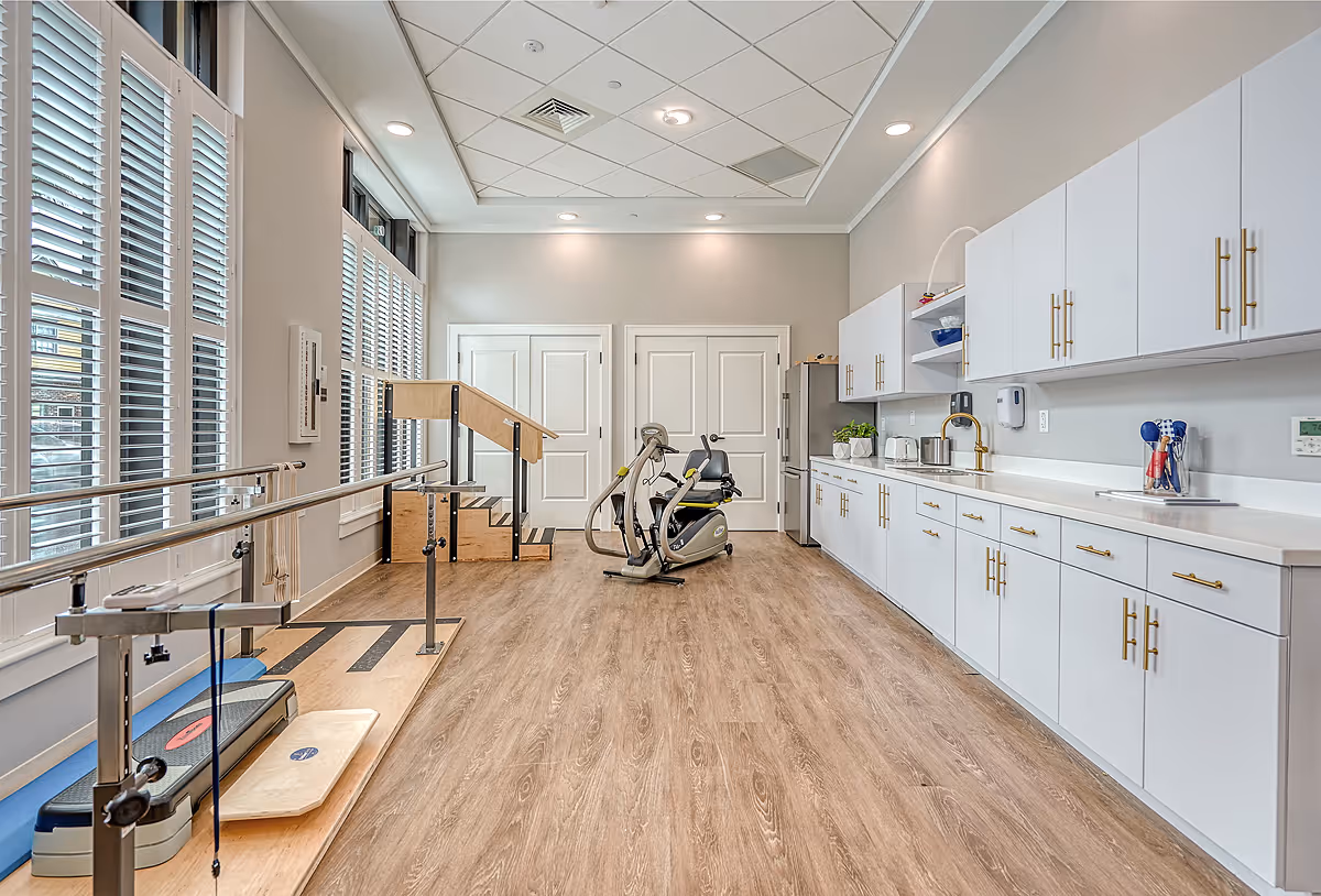 A bright rehabilitation room with large windows covered by white blinds on the left, wooden flooring, and white cabinets with gold handles on the right. The room contains physical therapy equipment including parallel bars, a balance board, a stationary exercise bike, and a set of stairs with handrails for mobility training.