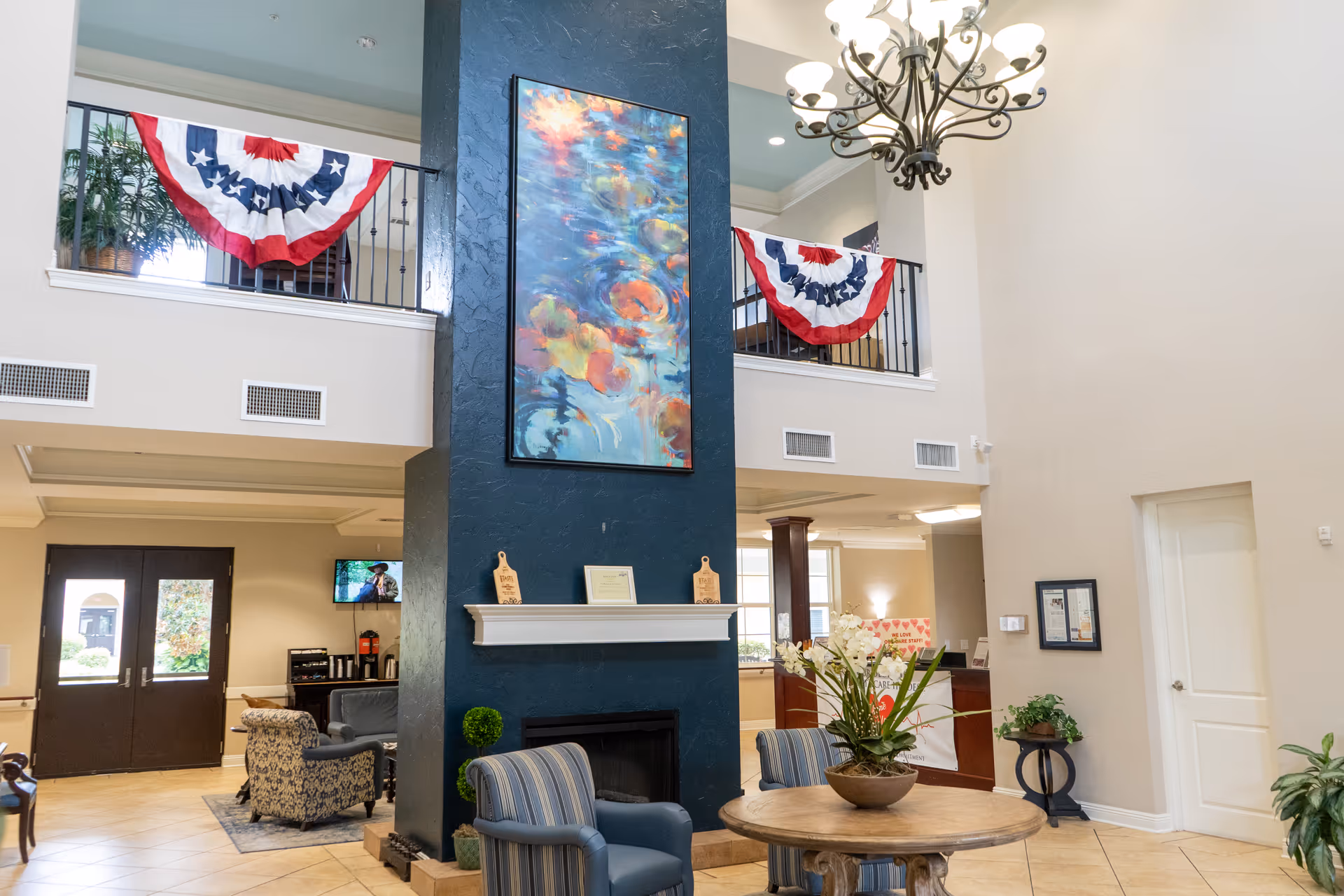 Interior view of a senior living facility lobby with a tall blue fireplace in the center, decorated with a colorful abstract painting. There are two patriotic red, white, and blue banners hanging from the upper balcony railing. The lobby has comfortable seating including striped armchairs around a round wooden table with a floral arrangement. A chandelier hangs from the ceiling, and there is a reception desk visible in the background.