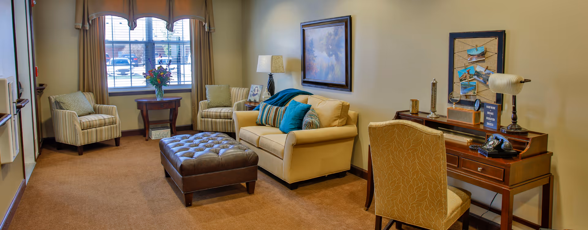 A cozy living room with a sofa and colorful pillows, two armchairs by a window, a tufted ottoman, and a wooden desk with a chair.