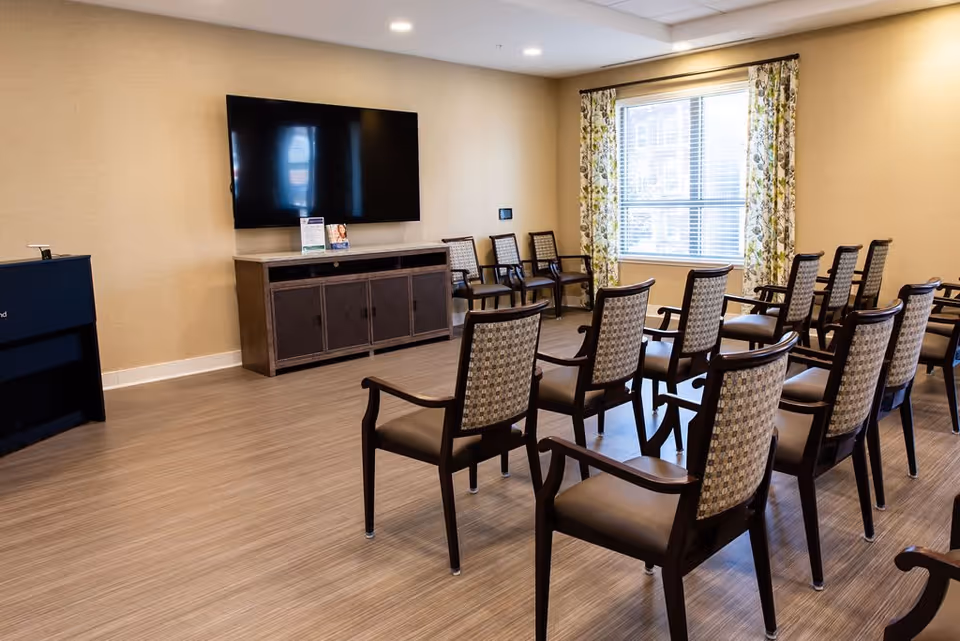 A meeting or presentation room with multiple rows of wooden chairs with patterned upholstery facing a large flat-screen TV mounted on a beige wall. There is a window with floral curtains letting in natural light, and a wooden cabinet beneath the TV. The floor is covered with light wood laminate.