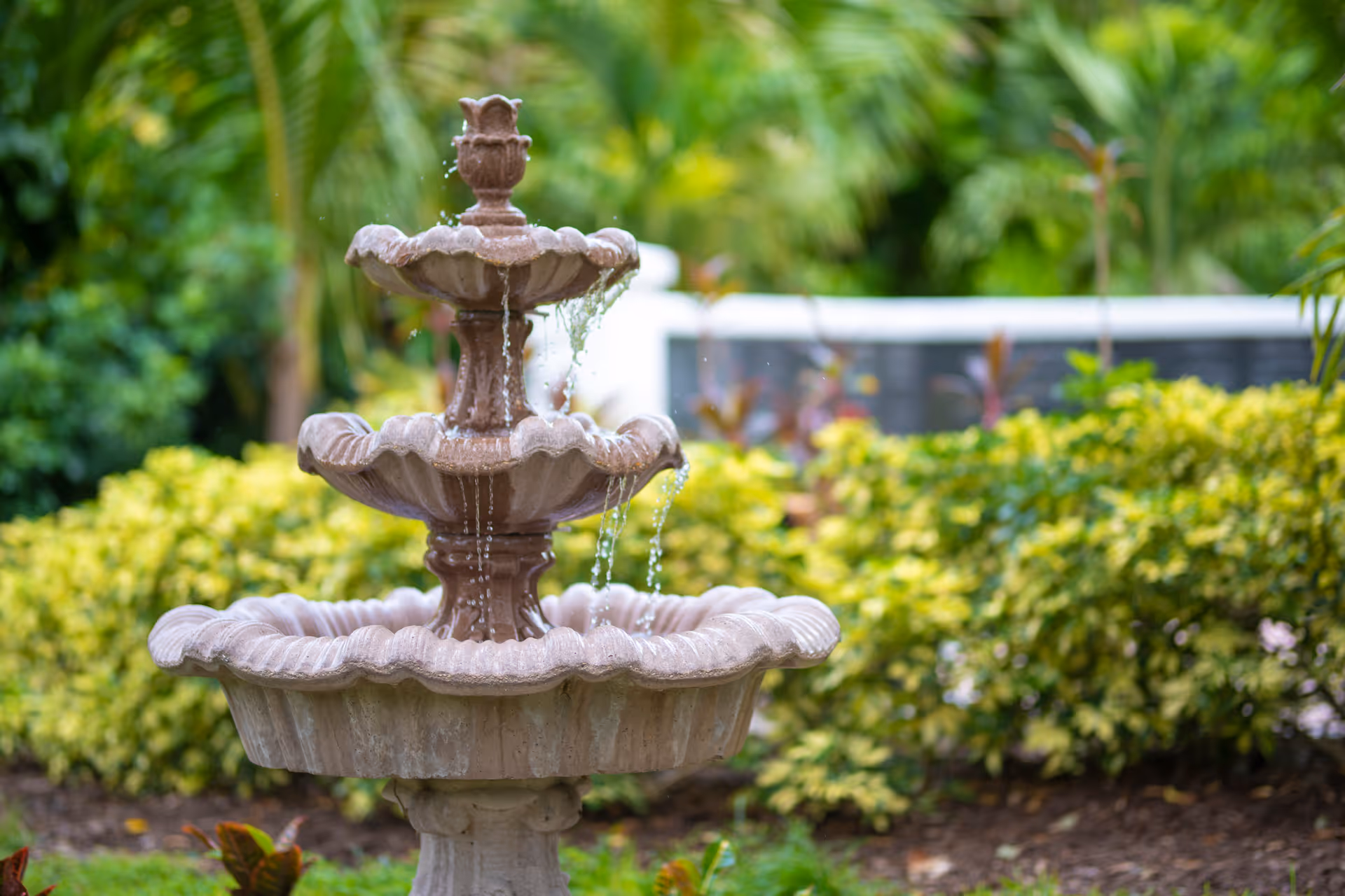 A three-tiered stone water fountain with water flowing from the top tier to the lower tiers, surrounded by green bushes and tropical plants in an outdoor garden setting.