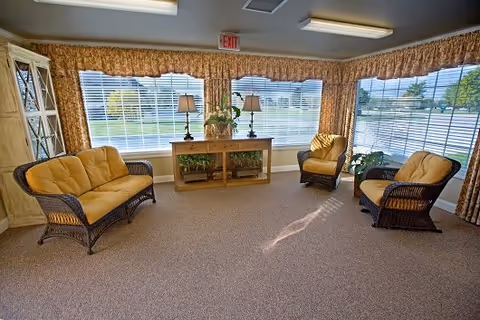 A bright living room area with large windows covered by blinds and floral valances. The room features a wicker loveseat and two wicker armchairs with yellow cushions arranged around a wooden console table with two lamps and a flower arrangement. The carpeted floor and the exit sign above the window are also visible.